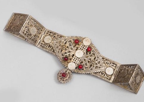 Ceinture en argent, Algérie, Aurès, XXe siècle.
Onze plaques articulées moulées dorées et ajourées de rosettes à c?ur de pièces de monnaie française. Fermoir en mandorle ajourée et aiguille sommée d'une rosace, à cabochons de verre rouge. 
Dim.: 40 cm
P.B.: 505 g
An articulated silver belt, Algeria, Aurès, 20th century