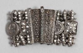 Bracelet, trois fibules et un pendant en argent, Algérie, XXe siècle. 
Bracelet à deux rangées de barrettes reliées par des chaînettes, encadrant une frise de médaillons inscrits « malbous al- Afiat» (apporte le bien-être) alternant avec des carrés étoilés, fermoir filigrané ; - Une paire et une fibule triangulaire à décor ajouré végétal, poinçon à tête de Maure ;- Un pendant triangulaire à cabochon de verre rouge, poinçon à tête de Maure.
Dim. bracelet: 7,5 x 4 cm ; Long. fibules: 6.5 cm, 5.5 cm ; Pendant: 4.5 cm
PB: 136 g
A silver bracelet, three silver fibulae and a silver pendant, Algeria, 20th century