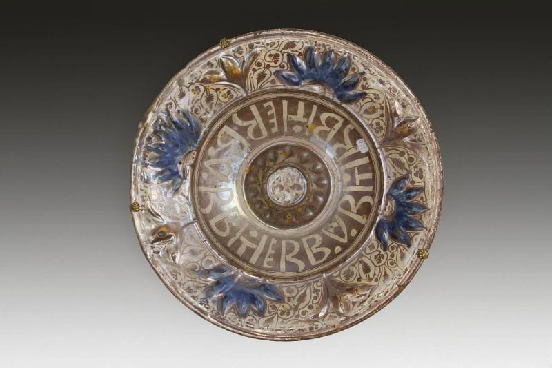 Plat lustré à ombilic, Espagne, Manises, XVIe siècle.
Plat creux à ombilic en faïence à décor de lustre métallique cuivré et rehauts de bleu sur fond crème. Inscription en latin sur le cavet. Au dos, décor de feuillage et lignes concentriques, trous de suspension. 
Diam.: 39,5 cm 
État: éclats 
A lustre dish, Spain, Manises, 16th century 