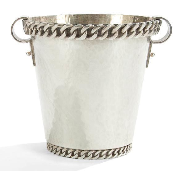 Jean DESPRES (1889-1980)

Très rare seau à rafraîchir moderniste, à deux gourmettes, en métal argenté à surface entièrement martelée.
La base en retrait et le pourtour du col sont au décor d'une épaisse gourmette à maillons plats en application.
Les anses, formées d'une large lame, décrivent un cercle tronqué se poursuivant par une languette fixée par une sphère.
Signé et daté à la pointe J. DESPRÉS 1965 sous la base.
Hauteur: 21,5 cm
Provenance:
- Madame Yvonne André (1905-1992) à Vézelay, ?uvre acquise directement auprès de l'artiste dans sa boutique d'Avallon.
- Par descendance, Madame Léone Danielle D. (née en 1924), fille de la précédente."