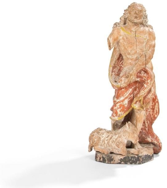 Saint Jean Baptiste en bois sculpté en ronde-bosse et polychromé. Debout, l'agneau à ses pieds, le corps ceint d'un manteau revenant sur le devant, il lève le regard vers le ciel; base monoxyle à pans. XVIIe siècle. H.: 50 cm.(fentes, manque le bras droit)