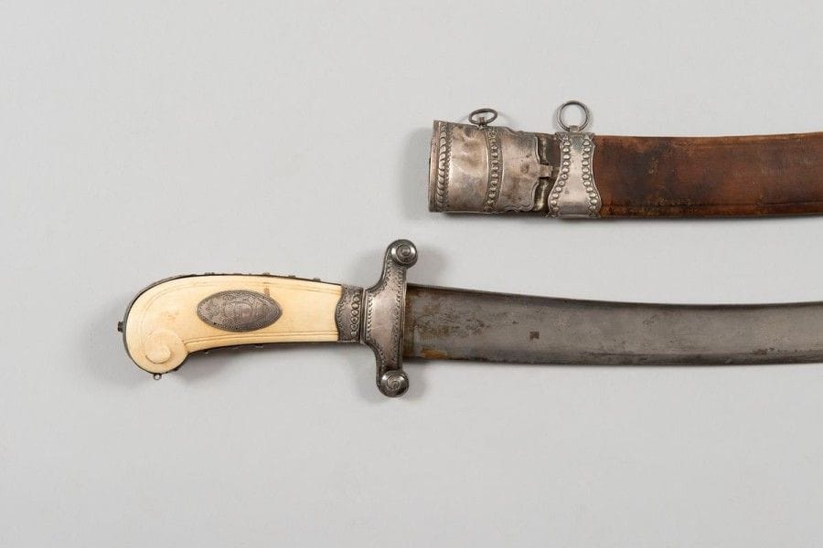 Couteau de vénerie. 
Poignée en ivoire ornée sur chaque face d'une plaque ovale en argent gravé, sur l'une, d'armoiries («d'argent à la fasce de sable à trois annelets, surchargé de trois roses») sous couronne de marquis et sur l'autre du monogramme «GB» dans une guirlande de feuillages et rubans. 
Monture et croisière en argent. Forte lame à dos plat, contre-tranchant et langue de carpe. Avec son fourreau en cuir brun (rétréci) à trois garnitures en argent décoré en suite. 
B.E. Vers 1780