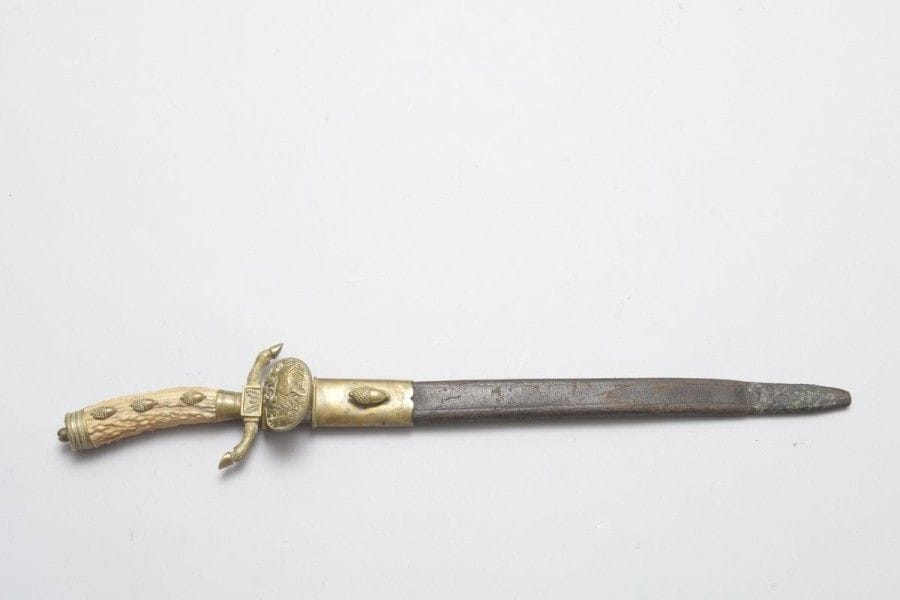 Couteau de vénerie allemand. 
Poignée en bois de cerf à trois boutons de rivure. Monture en laiton, clavier orné d'un cerf bramant. Lame droite à dos gravé (oxydations). Fourreau en cuir (rétréci) à deux garnitures en laiton. 
A.B.E. Fin XIXe siècle