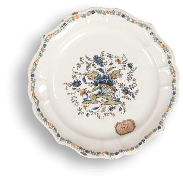 NOVE DI BASSANO

Assiette à bord contourné en faïence à décor polychrome au centre de rinceaux rocaille et branches fleuries sur terrasse.
XVIIIe siècle
D.: 23,5 cm
Egrenures
