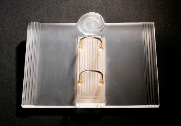 Maison LALIQUE
Joséphine Porte-agenda en métal doré et cristal moulé-pressé à décor de cannelures et d'un cygne. Un petit éclat. Signé LALIQUE ® France en lettres cursives à la pointe sur la tranche. 19,5 x 24,5 cm