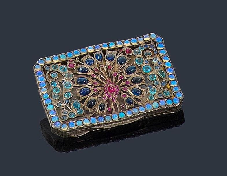 Boîte en argent, le couvercle ajouré d'un paon stylisé entre deux branches fleuries, l'ensemble serti de saphirs cabochons, d'aigues-marines, de rubis et de pâtes de verre, le fond extérieur gravé de rinceaux. Travail indien. Dimensions: 8,8 x 5,5 cm environ. Poids brut: 83,5 g