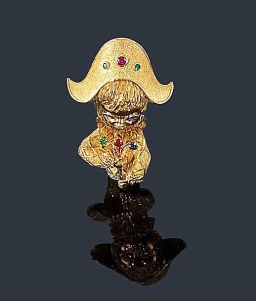 Broche-clip «Arlequin» sculpté en or jaune 18K (750 ?), le chapeau agrémenté de 2 émeraudes et d'un rubis, les yeux soulignés de saphirs, le bouquet piqué d'une émeraude, d'un rubis et d'un saphir, la ceinture agrémentée de diamants. Hauteur: 5,4 cm environ. Poids brut: 30,4 g