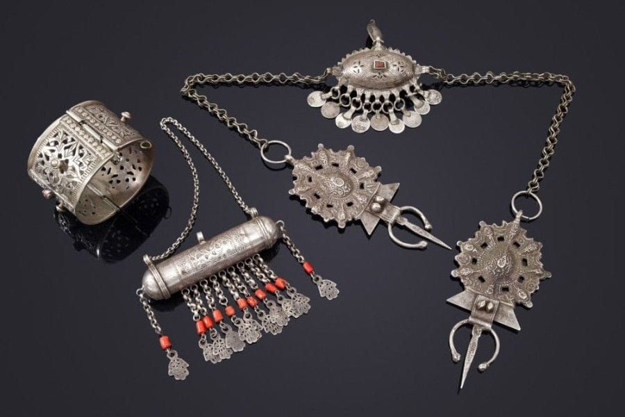 Porte - étui qannuta, parure pectorale kfrûn, et bracelet ajouré, Tunisie et Maroc, début XXe siècle
Porte - étui talismanique, qannuta cylindrique ciselé, à onze pendeloques, khamsa et perles de corail, Tunisie; parure pectorale de type tortue kfrûn, formée de deux fibules polylobées réunies à un médaillon centre en boule ajourée, à pendeloques de pièces du Maroc, région du Nador et Melila; un bracelet à charnière ajouré et incrustations de verre rouge.
A qannuta, a pectoral necklace kfrûn and a bracelet, Tunisia and Morocco, early 20th century