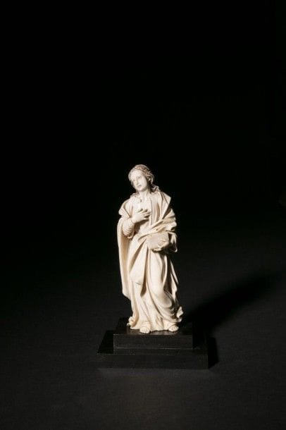 Saint-Jean en ivoire sculpté en ronde-bosse avec rehauts de polychromie. Debout, une main sur la poitrine, l'autre portant un livre, il est vêtu d'une tunique et d'un manteau dont un pan revient sur le devant. 
Espagne, XVII / XVIIIe siècle.
H.: 18,5 cm
Socle en bois noirci
