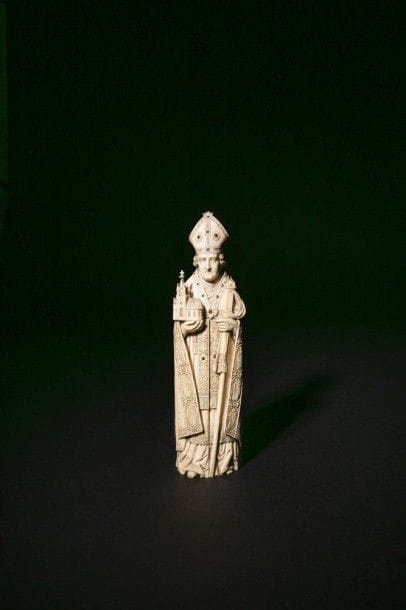 Saint Evêque fondateur en ivoire sculpté en ronde-bosse ouvrant et formant triptyque, incrustations de cabochons de verre de couleur. Debout, portant les attributs épiscopaux, il tient une église dans sa main droite; les deux vantaux s'ouvrent sur un retable avec plusieurs scènes: le Christ bénissant, la Crucifixion, l'Adoration des Mages, l'Ecce Homo, la Présentation au temple.
XIXe siècle.
H.: 31,4 cm