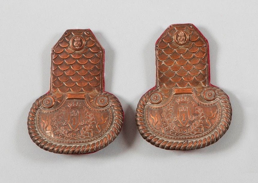 Paire d’épaulettes de musicien de la Garde royale, attribuée au 1er régiment. 
En cuivre, à décor d’écaille au chiffre du roi Louis XVIII, encadrant le chiffre « 1 ». Boutons au modèle de la Garde royale vers 1815-1816. 
Doublure en drap cramoisi. 
A.B.E. Vers 1815-1816.