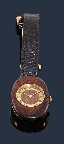 CARTIER.
Montre de dame en or jaune 18K (750‰) et bois d’amourette, cadran de forme ovale, index chiffres romains, bracelet en lézard à boucle déployante en or 18K, mouvement mécanique. Numérotée 1047. 
Longueur : 17,2 cm environ. 
Poids brut : 32,2 g 
(mouvement à réviser, manque le remontoir)