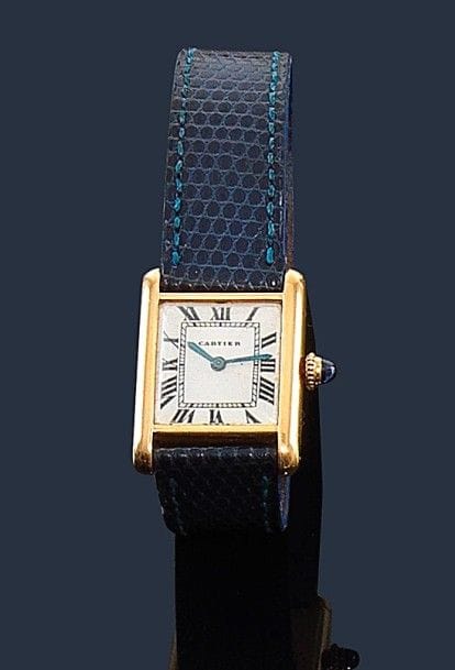 CARTIER.
Montre de dame en or jaune 18K (750‰), modèle Tank, fond blanc, index chiffres romains, remontoir saphir cabochon, bracelet en lézard bleu à boucle déployante en or 18K, mouvement mécanique. Numérotée 023979 52944. 
Longueur : 18 cm environ. 
Poids brut : 28 g 
(mouvement à réviser)
