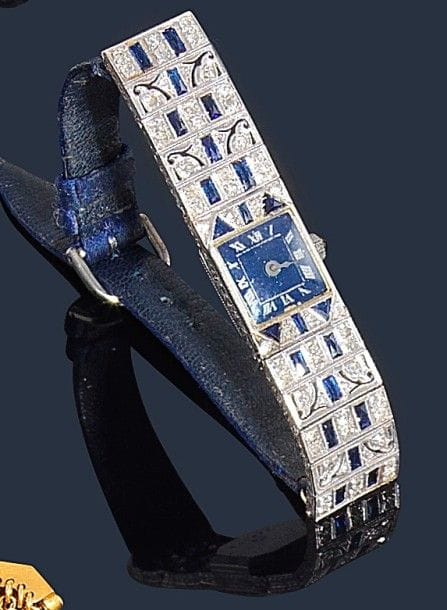Montre de dame en platine et or 18K (75‰), cadran carré, fond bleu, index chiffres romains, les attaches en platine ajouré, serti de diamants et de pierres bleues, bracelet tissu et boucle métal, mouvement mécanique. Travail des années 1930.
Poids brut : 19,7 g
(mouvement à réviser, manque une aiguille, bracelet rapporté)
