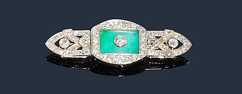 Broche en or gris 18K (750‰), ajourée, sertie de diamants de taille ancienne, ornée d’un jade.
Longueur : 5,5 cm environ.
Poids brut : 11,3 g (transformations)