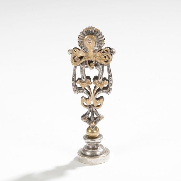 TRAVAIL ART NOUVEAU 
Précieux cachet en métal à patine argent et dorée, à décor d’un profil de femme à la chevelure abondante se mêlant à des rinceaux végétaux. 
H. : 8,3 cm