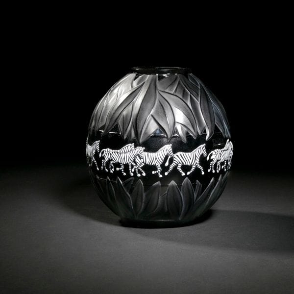 Maison LALIQUE d’après un modèle de Marie-Claude LALIQUE (1935-2003)
Tanzania, modèle créé en 1991
Vase. Épreuve en cristal noir à décor d’une frise tournante de zèbres, émaillée blanc et noir. (Lalique Catalogue Général 2012 page 133). Dans son coffret d’origine (abimé). Signé LALIQUE R France sous la base. H. : 21 cm 