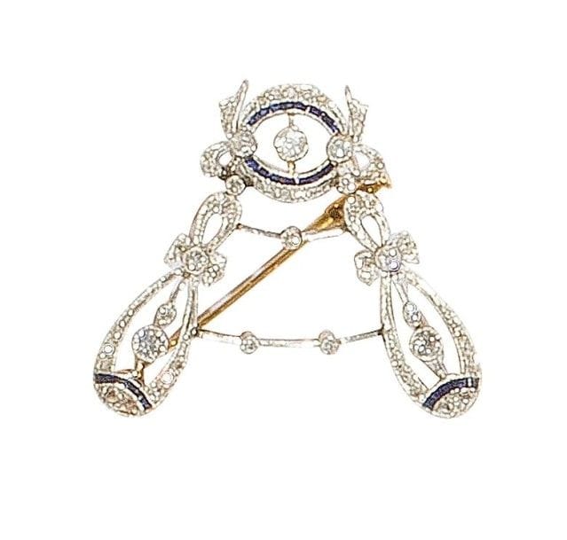 Broche en or gris et jaune 18K (750‰) formant un motif noué agrémenté de saphirs calibrés et de diamants taillés en roses terminé de deux motifs allongés sertis de diamants et de saphirs. 
Travail français du début du XXe siècle. 
Haut. : 3 cm environ. 
Poids brut : 6 g