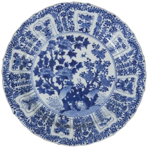 CHINE - Époque KANGXI (1662 - 1722)
Assiette en porcelaine décorée en bleu sous couverte d’un rocher percé et fleuri de pivoines, la chute ornée de réserves en forme de pétales et décorées de fleurs de tournesol. Au revers, la marque à la conque. (Fêlure). Diamètre : 24,5 cm