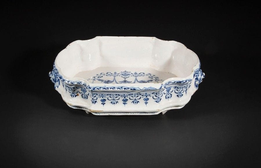 Moustiers 
Bassin ovale à bord contourné en faïence muni de deux prises latérales en forme de masque de faune à coiffe indienne, à décor en camaïeu bleu dans le style de Berain au centre d’un amour ailé sur une console cerné de sphinges, volatiles, draperies suspendues, guirlandes et rinceaux feuillagés.
Premier tiers du XVIIIe siècle. 
L : 41 cm, H : 11 cm
Quelques manques d’émail.