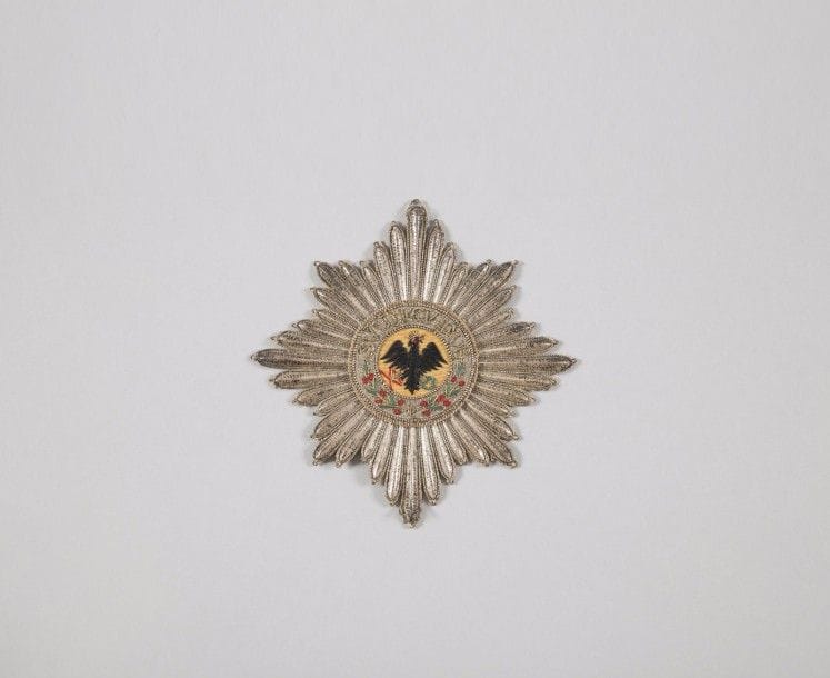 ALLEMAGNE (PRUSSE)
ORDRE DE l’AIGLE NOIR, fondé en 1701.
Grande plaque d’habit de Chevalier.
Étoile à huit branches rayonnantes en lamé, et fils d’argent.
Centre finement brodé sur fond de fil de moire orangée, d’un aigle couronné tenant un foudre et une couronne de lauriers, en fil noir, vert et rouge. Légende brodée en suite « SUUM CUIQUE » et branches de laurier.
Doublée d’origine en papier. Assez bon état (quelques fils décousus).
Diamètre : 13,1 cm
Première partie du XIXe siècle. 
Provenance : Famille Benoist-Mechin