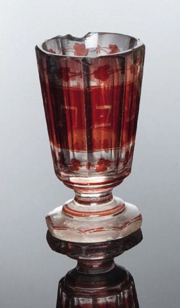 GOBELET DE KIDDOUSH Verre de Bohême à fond rouge, portant un décor peint d'inscription hébraïque et feuillages. Modèle à huit pas reposant sur un piédouche octogonal. (accidents et manques au bord supérieur et au pied). Haut.: 10,2 cm