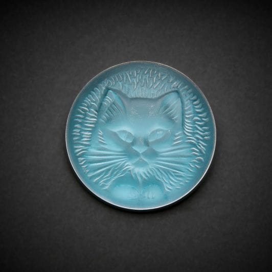 Maison LALIQUE d’après un modèle de Marie-Claude LALIQUE (1935-2003)
Tête de chat 
Broche en cristal moulé-pressé, achevé satiné, sur tain miroir bleu. Platine en métal argenté. Signée LALIQUE au revers. D. 4,8?cm
