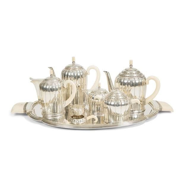 Paul FOLLOT (1877-1941) Décorateur & Henri Émile LAPPARRA (1891-1988) Orfèvre
 
Modèle présenté à l’Exposition des Arts Décoratifs et Industriels Modernes, Paris, 1925 
Service à thé et à café en argent composé de deux verseurs, un pot à lait, un pot à crème, un sucrier et un passe-thé. 
Les pièces légèrement balustres, les couvercles au décor de rangées d’olives, les anses et les prises en ivoire. 
Époque Art déco.
Fendillements épars sur les parties en ivoire. Chaque pièce marquée des poinçons de Maître Orfèvre et de garantie. 
Poids brut : 2.808,27 g
Vendu avec son grand plateau chantourné, et au modèle des verseurs, réalisé en métal argenté et ivoire. 
Désargenture partielle. 
L. 59,5 cm
Bibliographie :
L’orfèvrerie française au XXe siècle – Tony Bouilhet, éditions Émile-Paul Frères, Paris, 1937. Modèle identique reproduit dans cet ouvrage