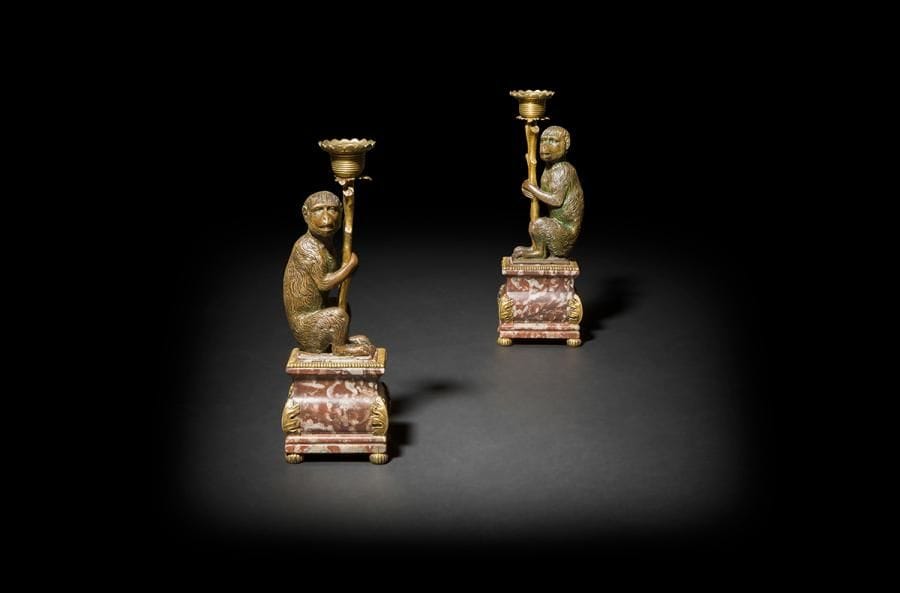 Rare paire de flambeaux aux singes porte-lumière, les figures de singe de profil tenant une branche d’arbre terminée par une bobèche à décor guilloché, reposant sur une base en marbre rouge ; (traces de laque sur les corps des singes).
Probablement Allemagne du sud, fin du XVIIe siècle.
H : 23 cm

Ce type de ciselure à larges traits imitant le pelage se retrouve sur certains bronzes réalisés à Augsbourg à la fin du XVIe siècle. Citons à cet égard notamment l’extraordinaire lièvre en bronze aujourd’hui conservé au Bayerisches Nationalmuseum à Munich (ill. in Bayerisches Nationalmuseum Bildfürer 1, Bronzeplastick, Munich, 1974, p. 43).

Une figure de singe jouant du violon réalisée à Augsbourg à la fin du XVIe siècle a été attribuée aux sculpteurs Hans Reisinger et Marx Labenwolf (Galerie Riudigier à Munich en 2016)
