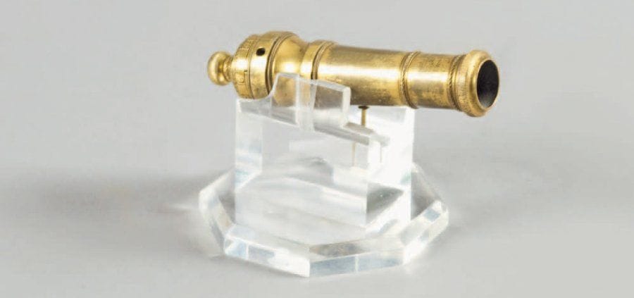 Curieuse et rare mesure à poudre. En forme de fût de canon, en bronze doré, à bourrelet à la bouche et anneaux de renfort. Bouchon de culasse à vis gradué de 1 à 12. T.B.E. Fin XVIIIe, début XIXe siècle. Présentée sur un socle en altuglas