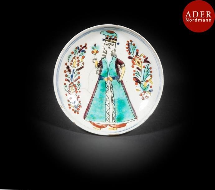 Coupelle à la jeune femme, Turquie ottomane, Kütahya, XVIIIe siècle 
Céramique siliceuse peinte à main levée en polychromie sur engobe blanc et sous glaçure transparente. Une jeune femme, portant une tunique turquoise ceinturée sur un salwar rouge et un turban assorti, tient une fleur en main droite. Des motifs composites floraux l’encadrent et deux lignes bleues au bord encerclent le tout. 
Diam. : 14,5 cm
État : quelques fêles et éclats aux bords.
A Kutahya pottery dish, Ottoman Turkey, 18th century