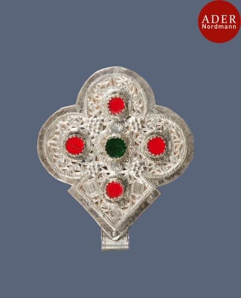 Amulette foulet khamsa, en argent, Maroc, Anti Atlas occidental, XXe siècle
En forme de trèfle, à décor repoussé et ciselé sur une face de rinceaux floraux ornés de cabochons de verre rouge et vert et sur l’autre de rosaces appliquées. Poinçons dont «1337».
Dim. : 12,6 x 14,7 cm ; P  : 139 g
A silver amulet, foulet khamsa, Morocco, Anti Atlas, 20th century 