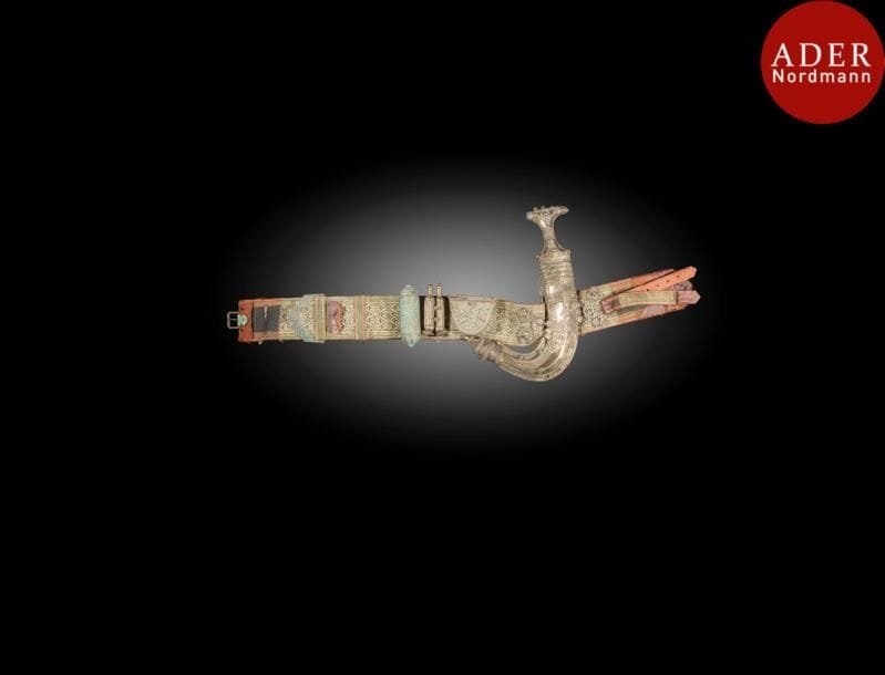 Poignard jambiya en argent, Yemen, XXe siècle 
Lame courbe en acier, poignée et fourreau à décor filigrané et ciselé géométrique et floral, monté sur une ceinture brodée de fils métalliques, agrémentée de deux boîtes talismaniques, cylindrique et demi circulaire, et d’un petit flacon en métal argenté.
Long. poignard : 35 cm ; long. ceinture 89 x 8cm ; PB  : 1376 g.
An Arabian silver horn-hiltted dagger, jambiyya, Yemen, 20th century