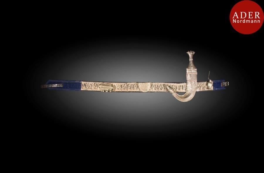Poignard jambiya en argent, Yemen, XXe siècle
Lame courbe en acier, poignée et fourreau à décor filigrané et ciselé de motifs géométriques floraux, fixé sur une ceinture brodée de fils métalliques, agrémentée de deux boîtes talismaniques, cylindrique et demi circulaire, et d’un petit flacon en métal.
Long. poignard : 31 cm ; dim. ceinture : 6 cm x 110 cm ; PB  : 806 g 
An Arabian silver horn-hilted dagger, jambiya, Yemen, 20th century 