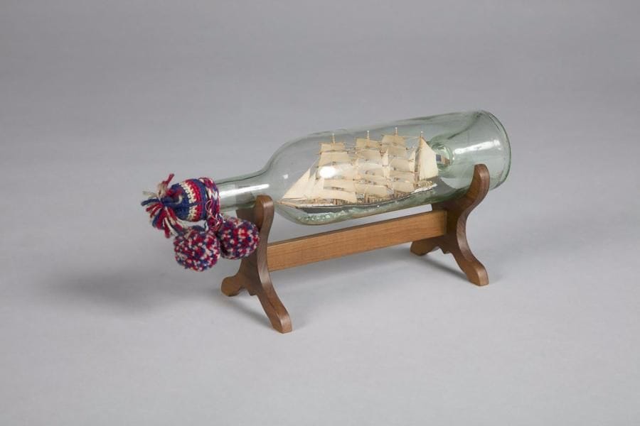 Voilier trois mâts en bouteille, toutes voiles montées. 
Bois sculpté peint. Bouchon tricoté en laiton tricolore, avec deux pompons. 
B.E. Sur socle. 
XXe siècle.