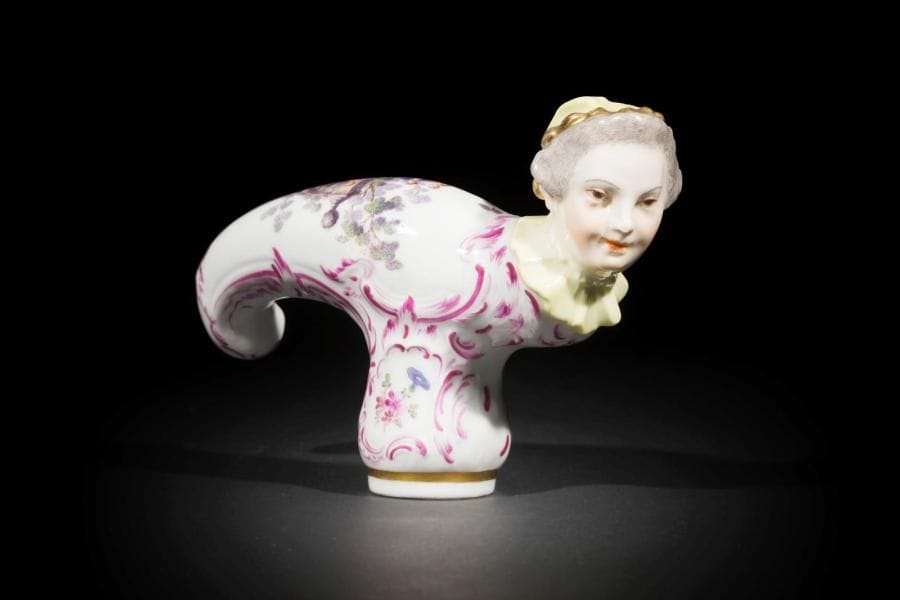 Meissen (genre de)
Pommeau de canne en porcelaine terminé par un visage de femme, à décor polychrome de couples galants dans un par cet bouquets de fleurs dans des encadrements de rinceaux rocaille en camaïeu pourpre.
XIXe siècle. 
L. : 12 cm