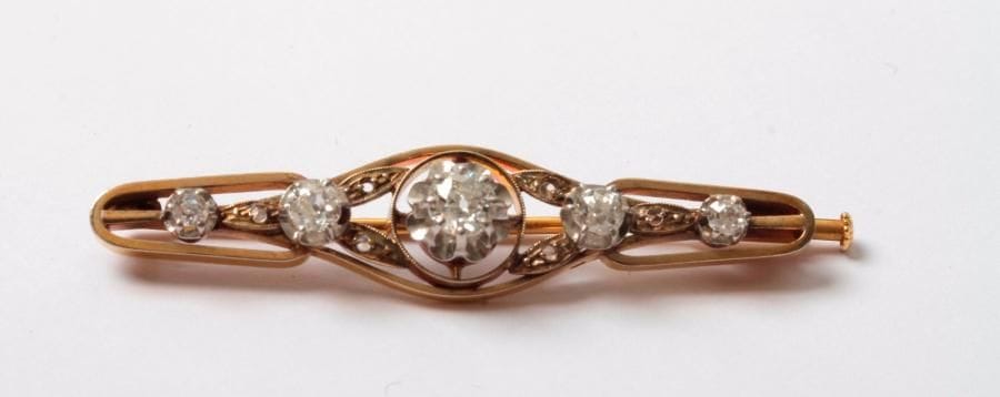 Broche en or 18K (750), ajourée, sertie d'une ligne de 5 diamants de taille ancienne, agrémentée de diamants taillés en roses. Longueur: 4,8 cm environ. Poids brut: 6 g (manque, chocs)