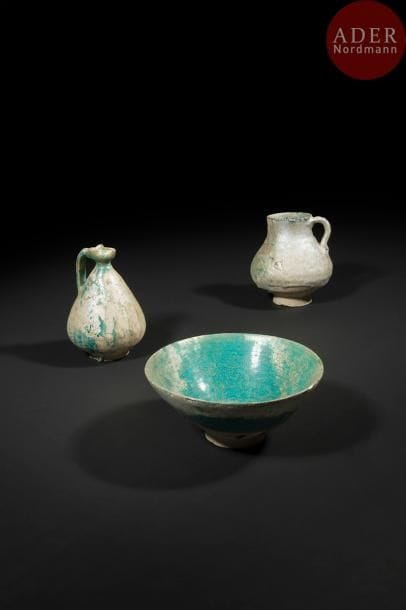 Trois céramiques turquoises, Iran XIIe - XIIIe siècle
Deux pichets en céramique siliceuse à glaçure turquoise, l’un à panse conique légèrement godronnée, et à bec tréflé avec anse ; l’autre à panse globulaire, col droit et anse en esse ; une coupelle à bords évasés sur base circulaire et traces d’incisions sur la panse. 
Haut. pichets : 6 cm, 5,4 cm ; diam. coupelle : 7,5 cm, haut. : 8 cm 
État : fortes irisations, manque au bord pour la coupelle et à la base pour les pichets. 
Two blue glazed pottery jugs and a bowl, Iran, 12th - 13th century.