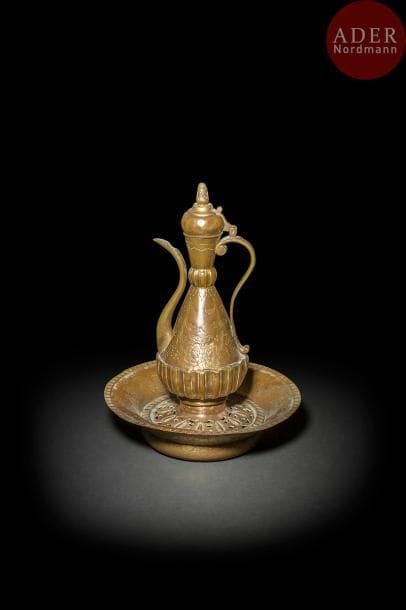 Aiguière, bassin et porte-savon en cuivre doré, tombak, Turquie ottomane, fin XVIIIe - déb. XIXe siècle
Aiguière à panse piriforme à col annelé à godrons et couvercle en dôme à prise en forme de pomme de pin. Décor ciselé de tiges florales tête-bêche sur un fleuron folié. Base goudronnée.
Porte-savon, sabunlunk, à décor ajouré ceinturé d’une frise ciselée de compartiments.
Le bassin reprend le même décor sur les bords.
Haut. aiguière : 33 cm. Diam. Bassin : 29,4 cm
État : dorure en partie effacée sur le bassin.
An Ottoman gilt-copper, tombak, ewer and matching basin, Turkey, late 18th- early 19th century.