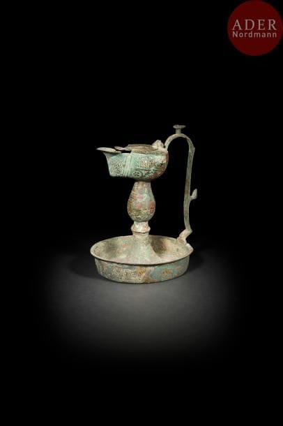Lampe à l’huile sur présentoir, Iran, XIIe siècle
Lampe ovoïde composite en bronze, à large bec avec couvercle, remontée sur un fût à bulbe et scellé sur une coupelle. Anse latérale surmontée d’une figurine. À décor gravé sur chaque partie de cartouches coufiques alternés de médaillons fleuris. 
H. : 20,7 cm
État : patine brune et verte, trou sur la lampe, composite de trois parties de différents objets. 
A composite oil lamp, Iran, 12th century. 