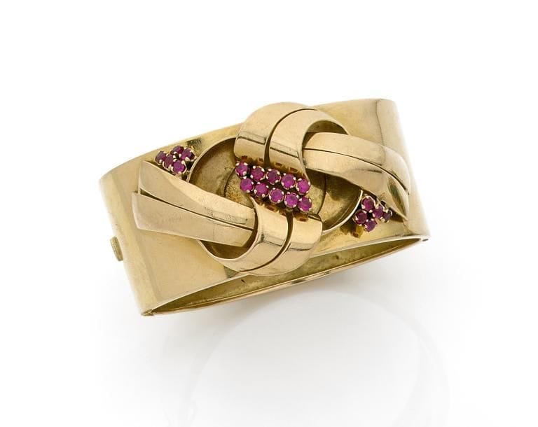 Bracelet manchette en or jaune 18K (750 ‰), appliqué d’un motif noué serti de rubis synthétiques. Travail des années 1940. 
Dimensions : 2,7 x 18 cm environ.
Poids brut : 67,5 g