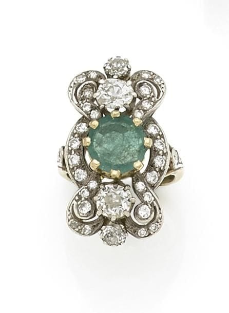Bague en or 18K (750 ‰) et argent, ornée d’une émeraude ronde entre deux diamants de taille ancienne, la monture formant des volutes serties de diamants. Travail français. 
Tour de doigt : 51.
Poids brut : 10,1 g