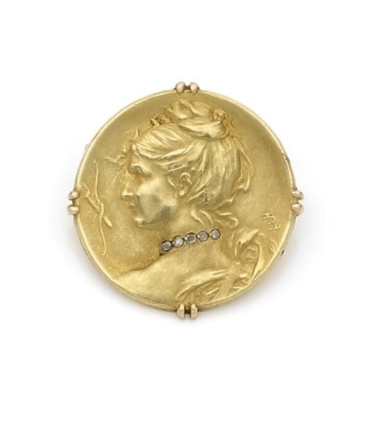 Broche en or 18K (750 ‰) ciselée d’une femme de profil portant les attributs de Diane chasseresse, le cou souligné d’une ligne de roses. Monogrammée LR. Travail français des années 1900. 
Diamètre : 2,4 cm environ.
Poids brut : 5,4 g