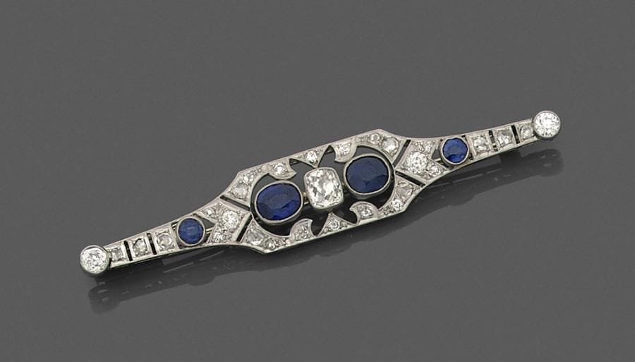 Broche fuseau en or gris 18K (750 ‰) et platine repercé de motifs géométriques imbriqués, ornée d’un diamant coussin de taille ancienne épaulé de saphirs ovales (dont deux synthétiques) et de diamants. Travail français des années 1925. 
Longueur : 7,5 cm environ.
Poids brut : 8,8 g