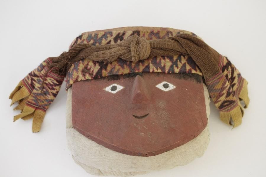 Fardeaux funéraire constitué d'un sac rembourré en tissu orné sur le dessus d'un masque humain au visage peint en rouge. Tissu, fibres végétales et bois polychrome. Culture Chancay, Pérou, 1100 - 1450 apr. J.-C. H. 28 - l. 27 - Ep. 19 cm. Un certificat d'Edouard Wagner Talleux sera remis à l'acquéreur.
