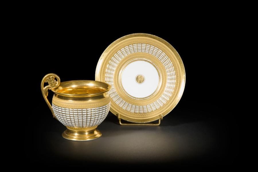 Grande tasse à chocolat et sa soucoupe en porcelaine à décor en relief rehaussé d’or de motifs imitant la vannerie et galons d’entrelacs à la molette à fond d’or laissé mat, l’anse élevée en forme de crosse, l’intérieur de la tasse à fond or.
Sans marque, probablement manufacture de Nast.
Début du XIXe siècle. 
H. 12 cm, D. 17 cm