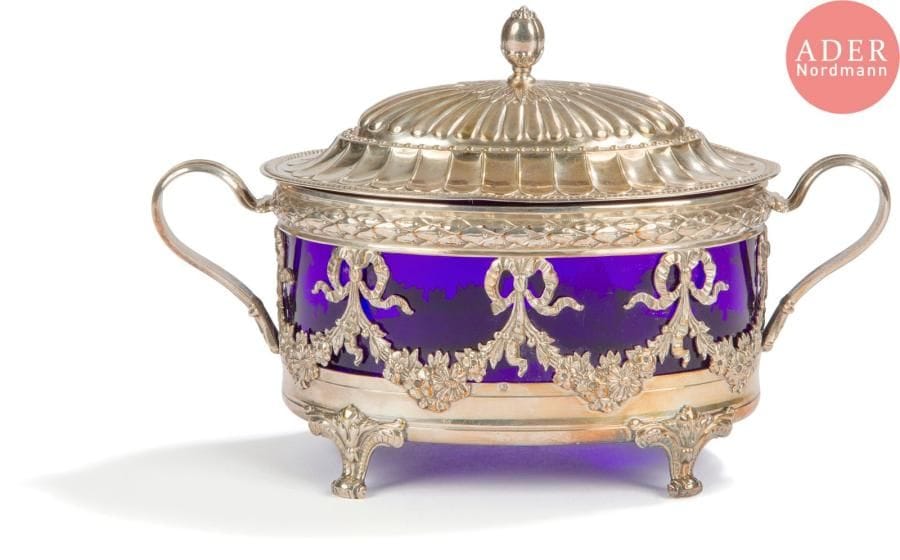 Sucrier en métal et son verre bleu de style Louis XVI. De forme ovale, quadripode, il est estampé de nœuds de rubans, repoussé de tors laurés, godrons et côtes torses sur le couvercle.
Travail étranger
Poids net : 265,60 g - longueur : 19,2 cm - largeur : 9,7 cm