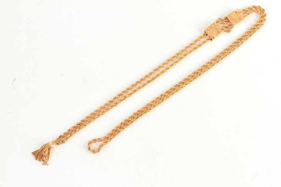 Long collier torsadé en or 18K (750 ‰) ponctué de deux motifs carrés ornés de fleurs, terminé par un pompon. Longueur : 120 cm environ. 
Poids : 58,3 g