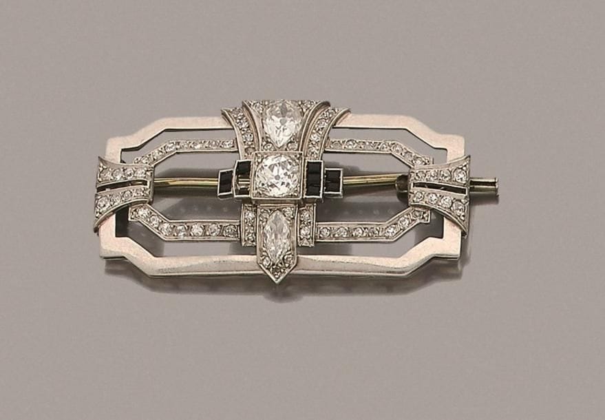 Broche-plaque en platine et en or gris 18K (750 ‰), ajourée de motifs géométriques imbriqués, centrée d’une ligne de trois diamants de taille ancienne, le premier taillé en goutte, le second rond, le dernier navette, épaulés d’onyx calibrés, sertie de diamants ronds de taille ancienne. Travail français des années 1930. Dimensions : 5 x 2,5 cm environ. Poids brut : 14,3 g (manquent deux onyx)