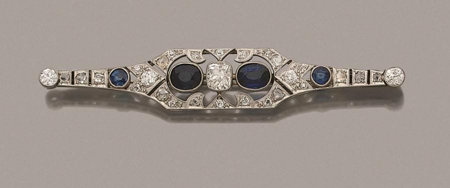 Broche fuseau en or gris 18K (750 ‰) et platine repercé de motifs géométriques imbriqués, ornée d’un diamant coussin de taille ancienne épaulé de saphirs ovales (dont deux synthétiques) et de diamants. Travail français des années 1925. 
Longueur : 7,5 cm environ.
Poids brut : 8,8 g